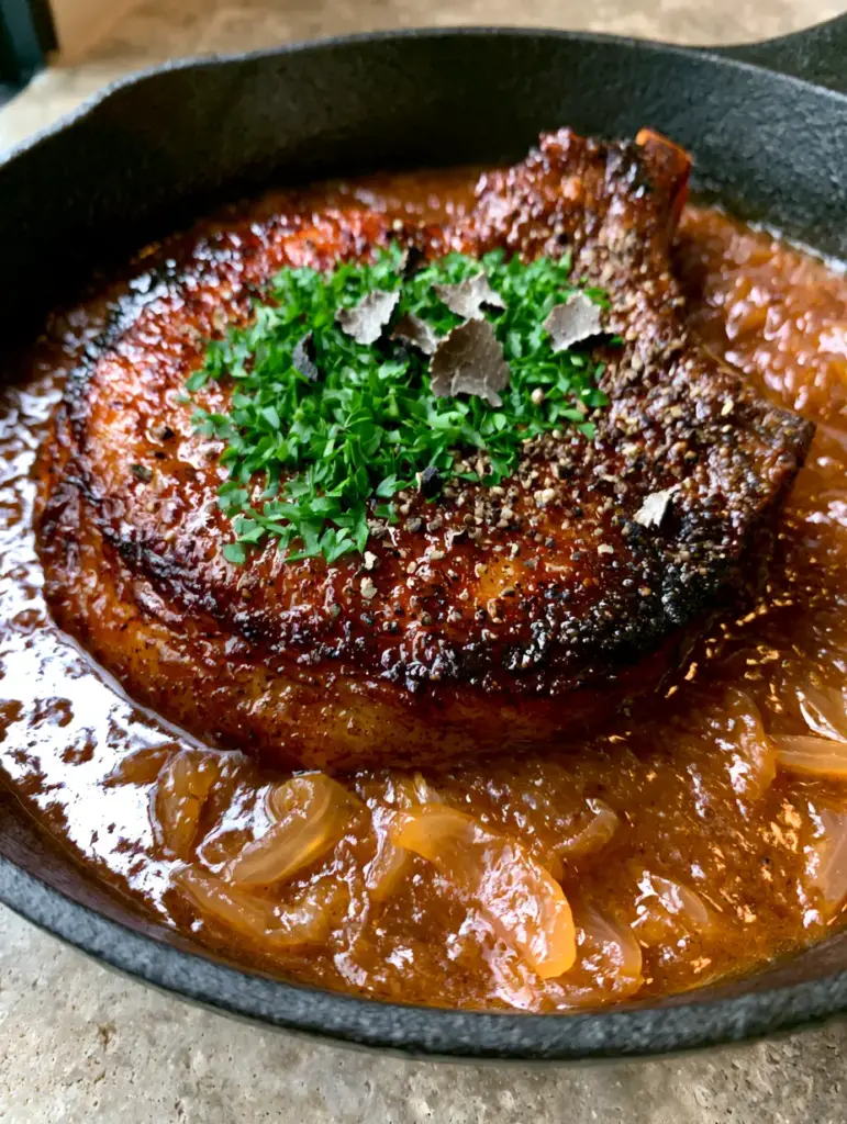 A top-down view of a single pork chop garnished with parsley and cracked black pepper in a mahogany onion sauce.